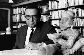 Arquitecto Pedro Ramírez sentado al interior de una biblioteca junto a una figurilla prehispánica